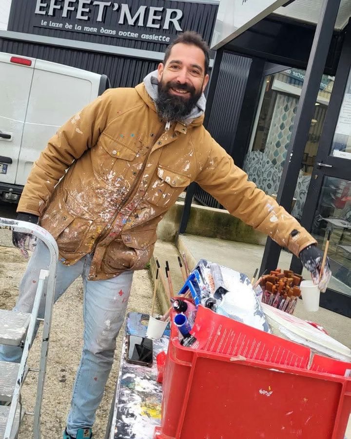Raphaël, souriant, devant son matériel de peinture, en veste tachée de peinture