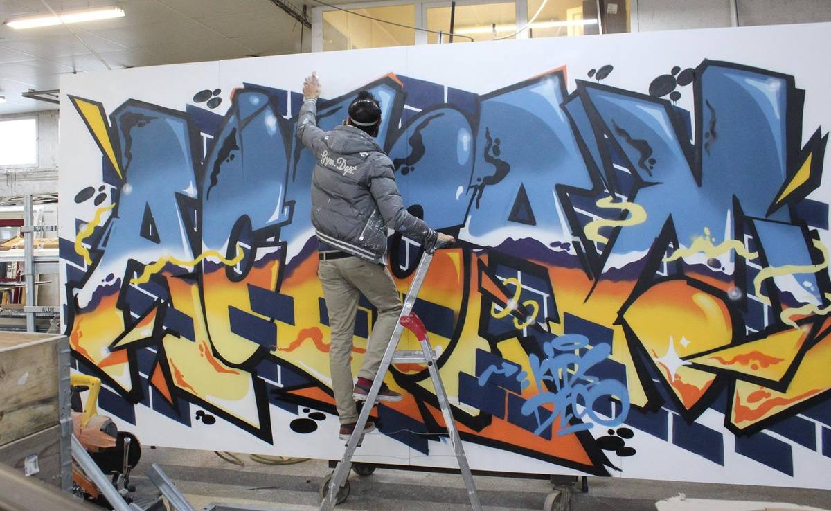 Raphaël peignant à la bombe un grand graffiti aux lettres bleues et orange sur un panneau, dans l'atelier d'une menuiserie