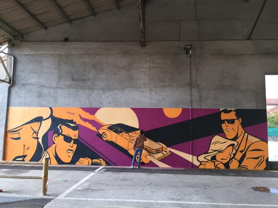 Grande fresque aux couleurs orange et violet dans le parking SNCF d'Ambérieu-en-Bugey : personnages de style bande dessinée rétro, voiture ancienne et soleil, avec Raphaël devant le mur