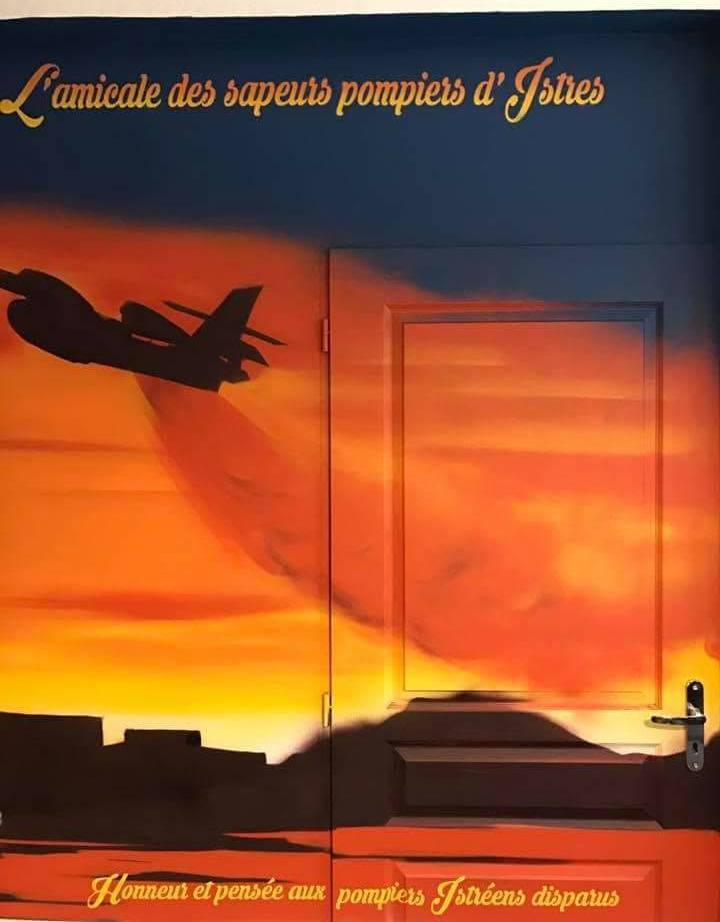 Fresque de l'amicale des sapeurs-pompiers d'Istres : avion bombardier d'eau sur un coucher de soleil, peint jusque sur la porte, « Honneur et pensée aux pompiers istréens disparus »