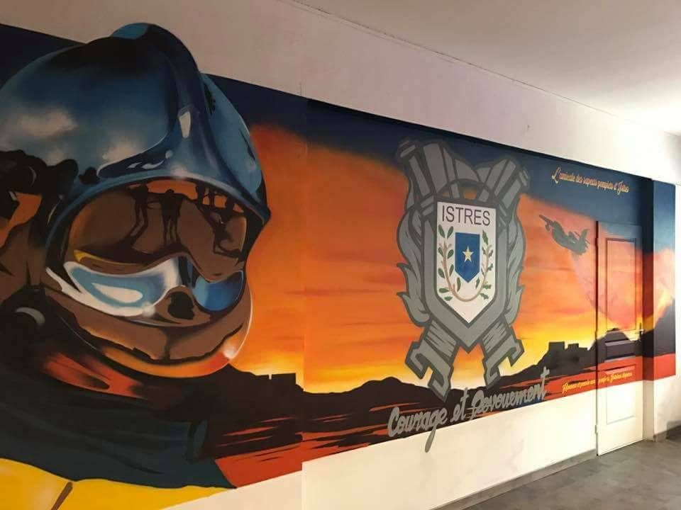Fresque dans un couloir de la caserne des pompiers d'Istres : casque de sapeur-pompier en gros plan, blason « ISTRES » et devise « Courage et Dévouement » sur un coucher de soleil orange, un avion bombardier d'eau au loin