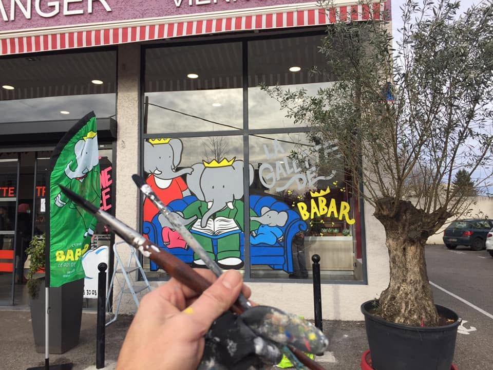 Vitrine de boulangerie « La galette de Babar » : la famille Babar lisant sur un canapé, pinceaux au premier plan