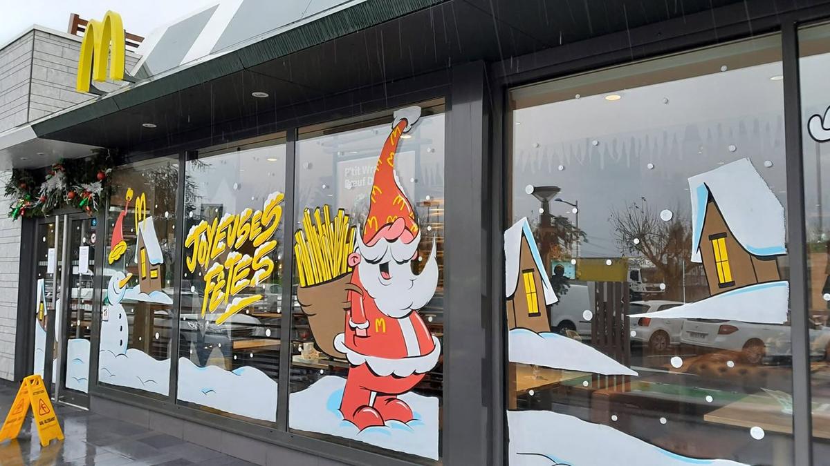 Vitrine de Noël peinte chez McDonald's : Père Noël portant une hotte de frites, lettrage « Joyeuses fêtes », bonhomme de neige et maisons enneigées