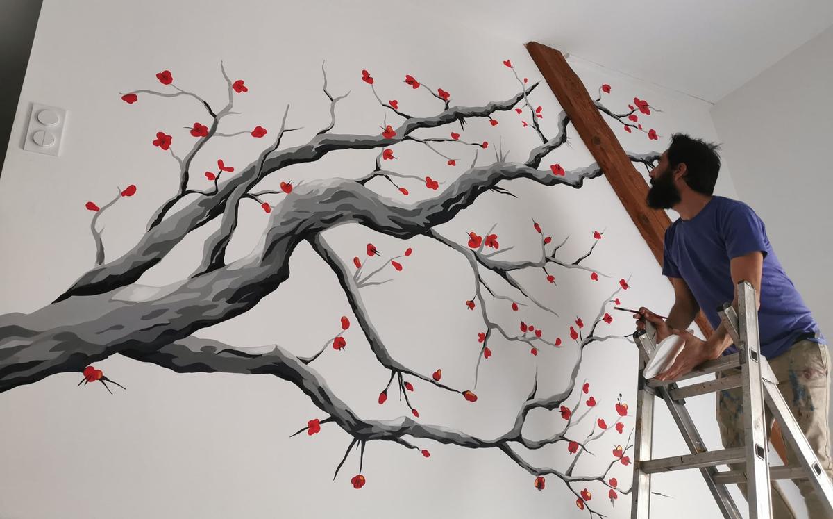 Raphaël sur son escabeau peignant une grande branche fleurie grise aux fleurs rouges sur un mur blanc, près d'une poutre en bois