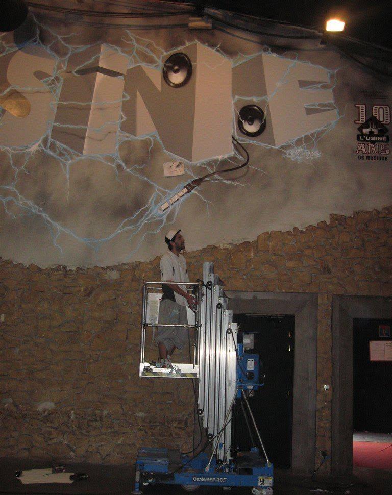 Raphaël sur une nacelle devant la grande fresque « L'Usine » avec lettres en relief, enceintes et éclairs, pour les 10 ans de la salle de concert